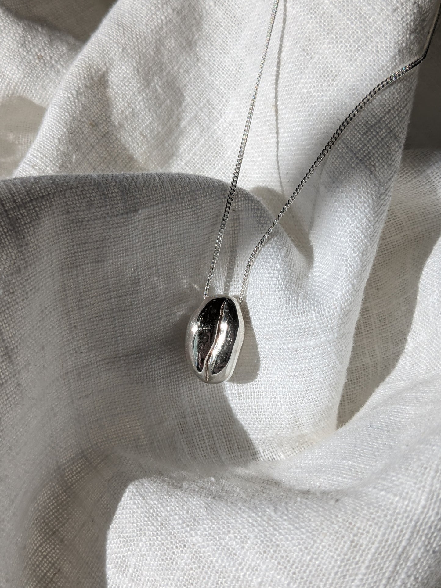 A silver coffee bean necklace laid out on a white fabric surface.