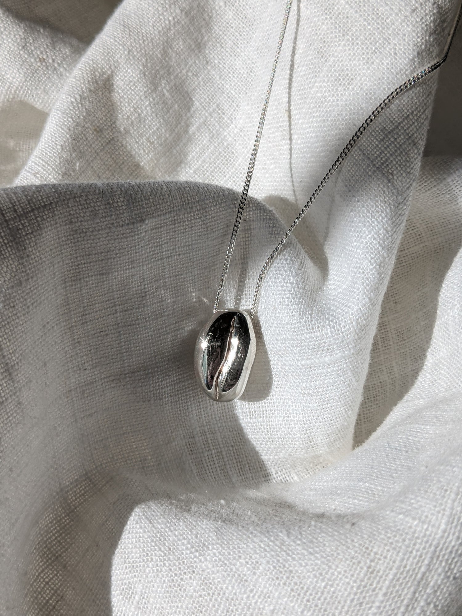 A silver coffee bean necklace laid out on a white fabric surface.