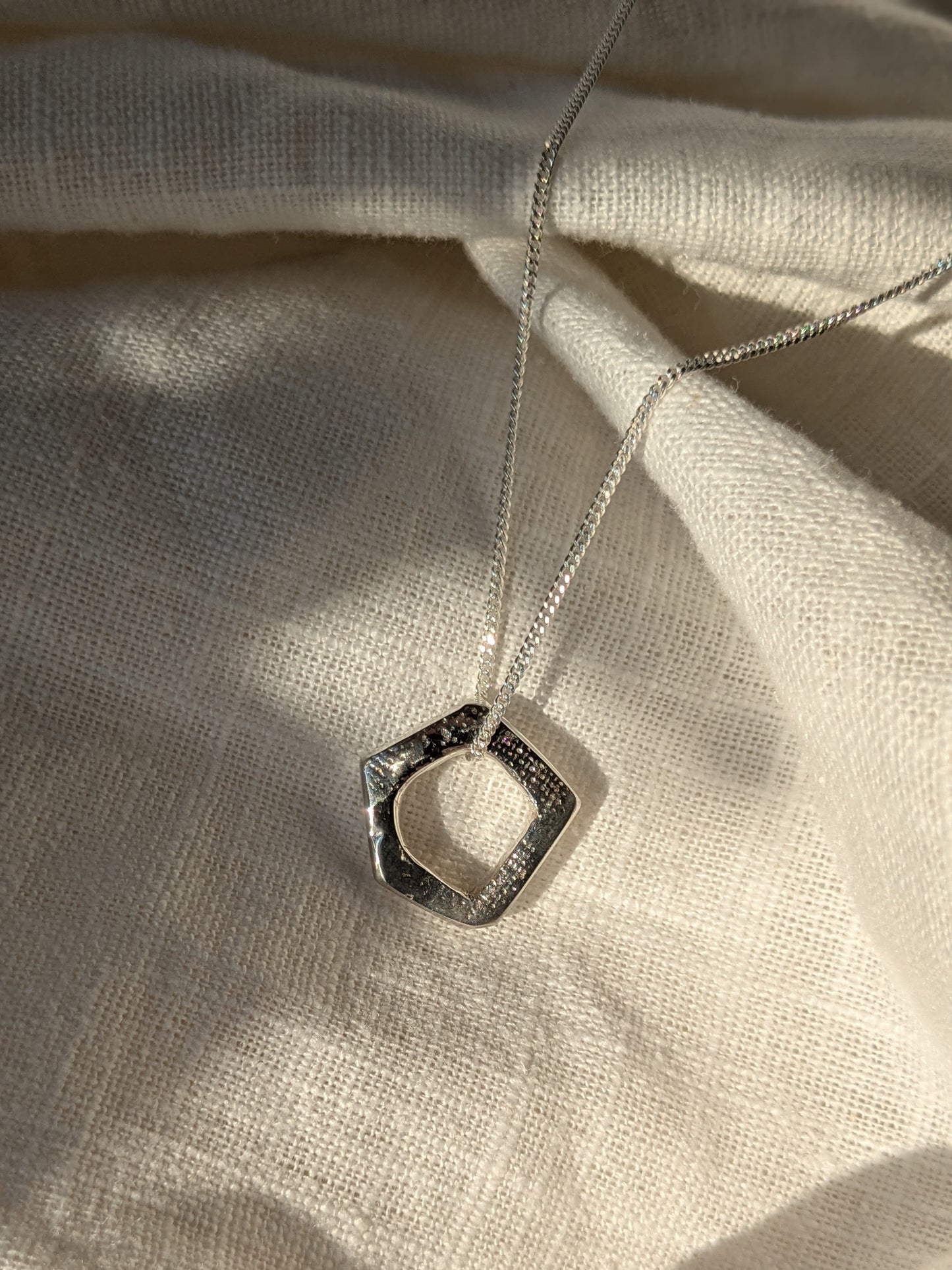 A silver pentagon-shaped pendant necklace with a textured irish linen surface on a white linen background.