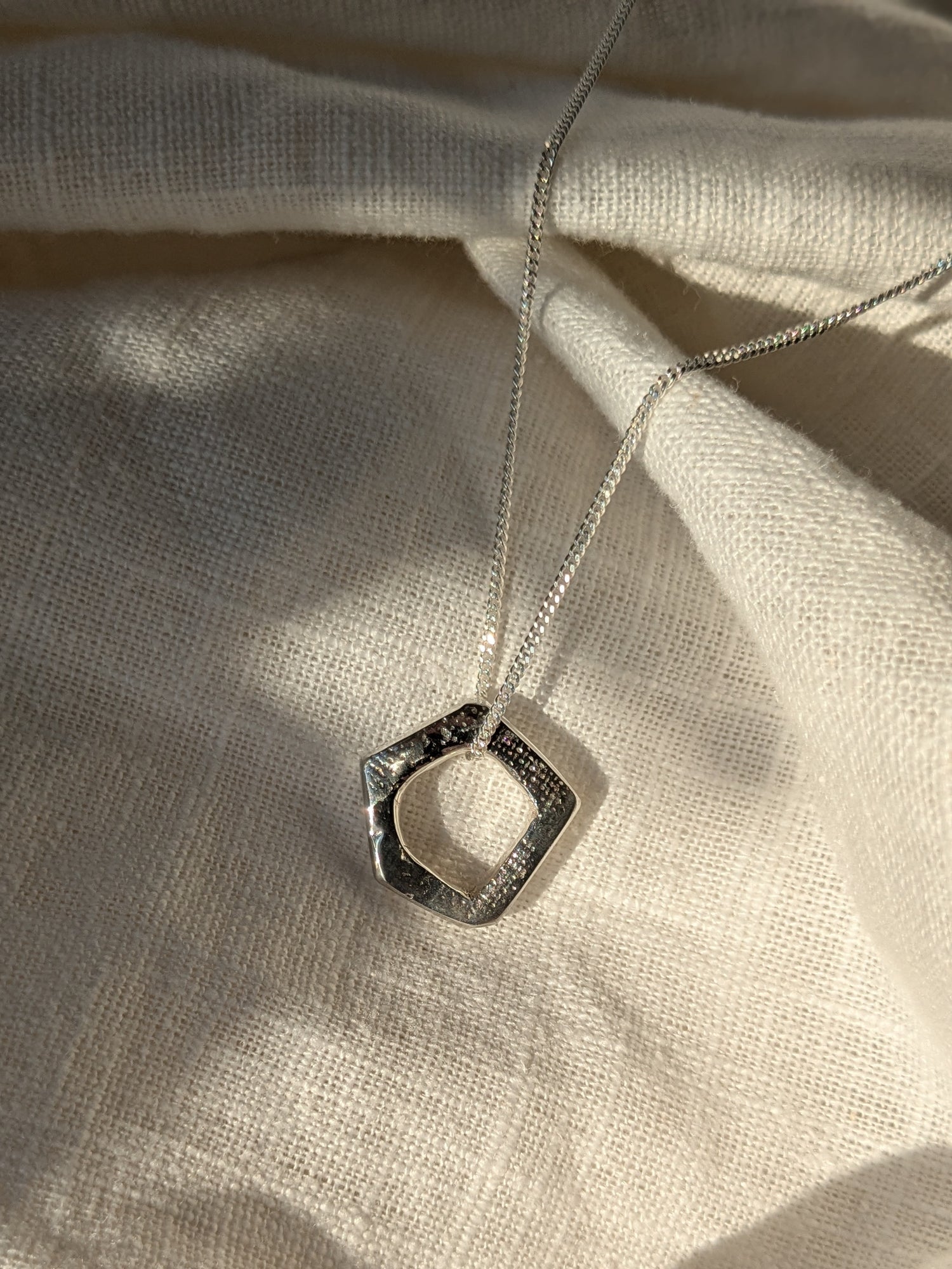 A silver pentagon-shaped pendant necklace with a textured irish linen surface on a white linen background.