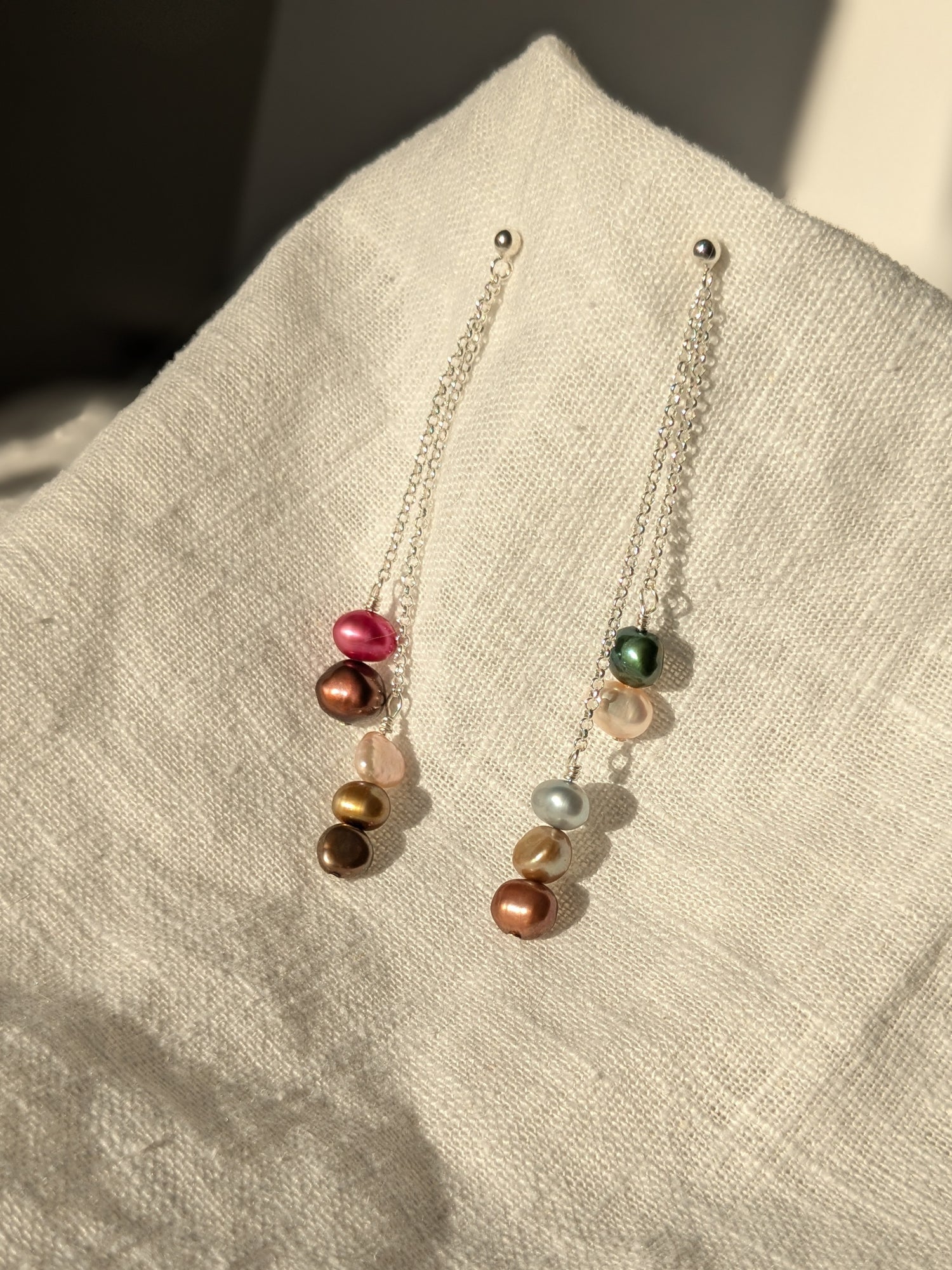 A pair of multi-colored pearl drop chain stud earrings laid on a textured fabric surface.