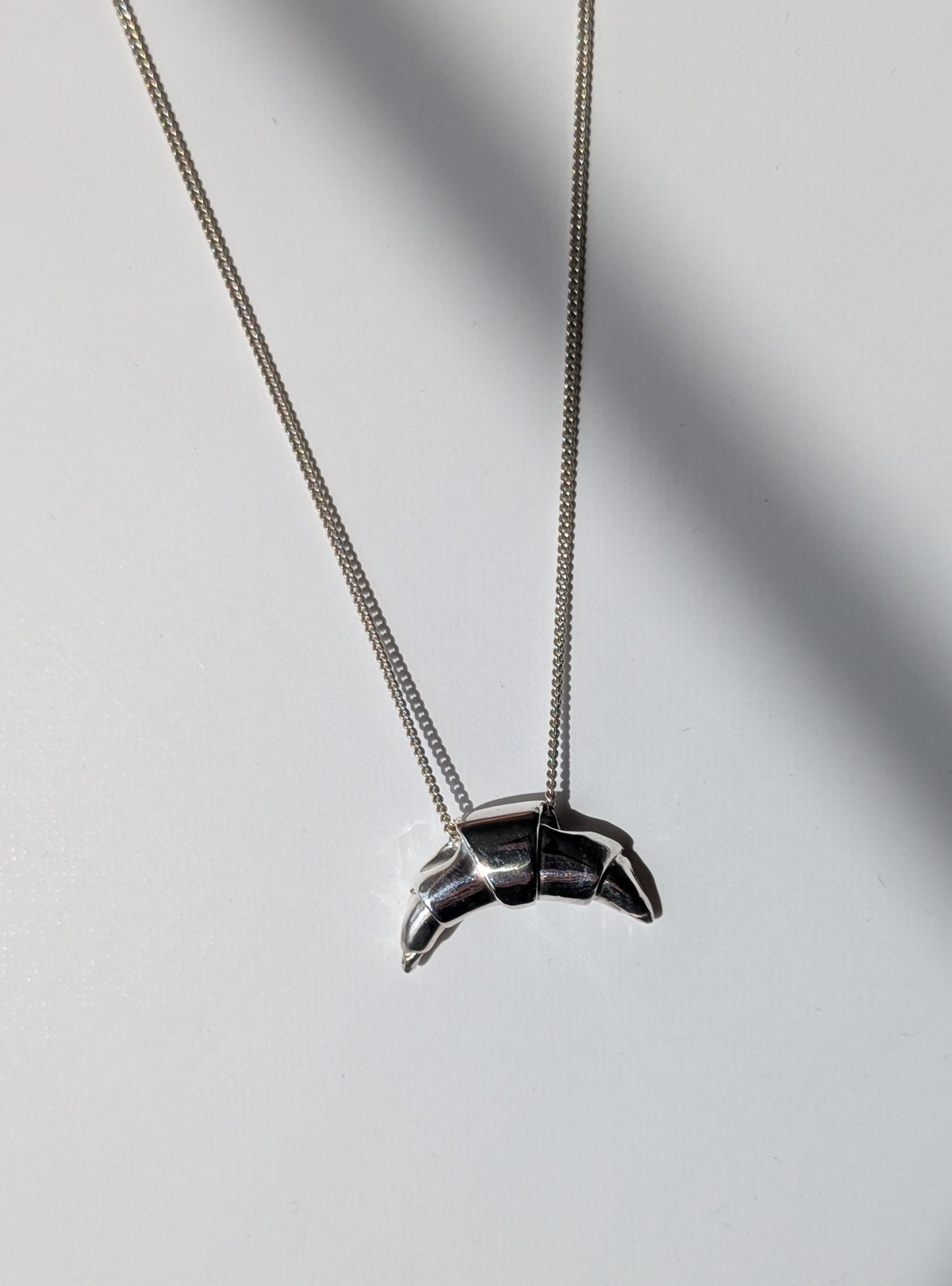 A silver croissant-shaped necklace pendant hanging on a chain, with a source of light creating a shadow and highlight on the pendant.