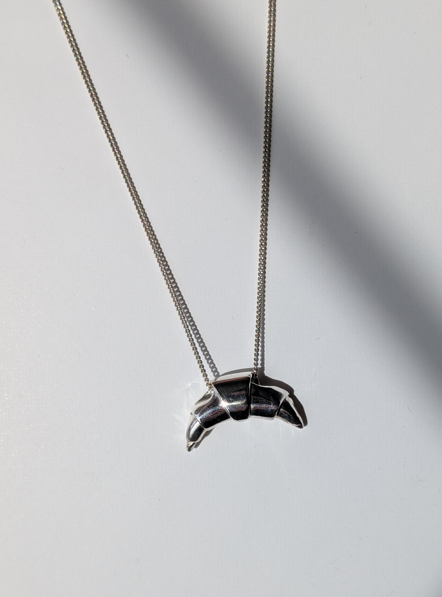 A silver croissant-shaped necklace pendant hanging on a chain, with a source of light creating a shadow and highlight on the pendant.