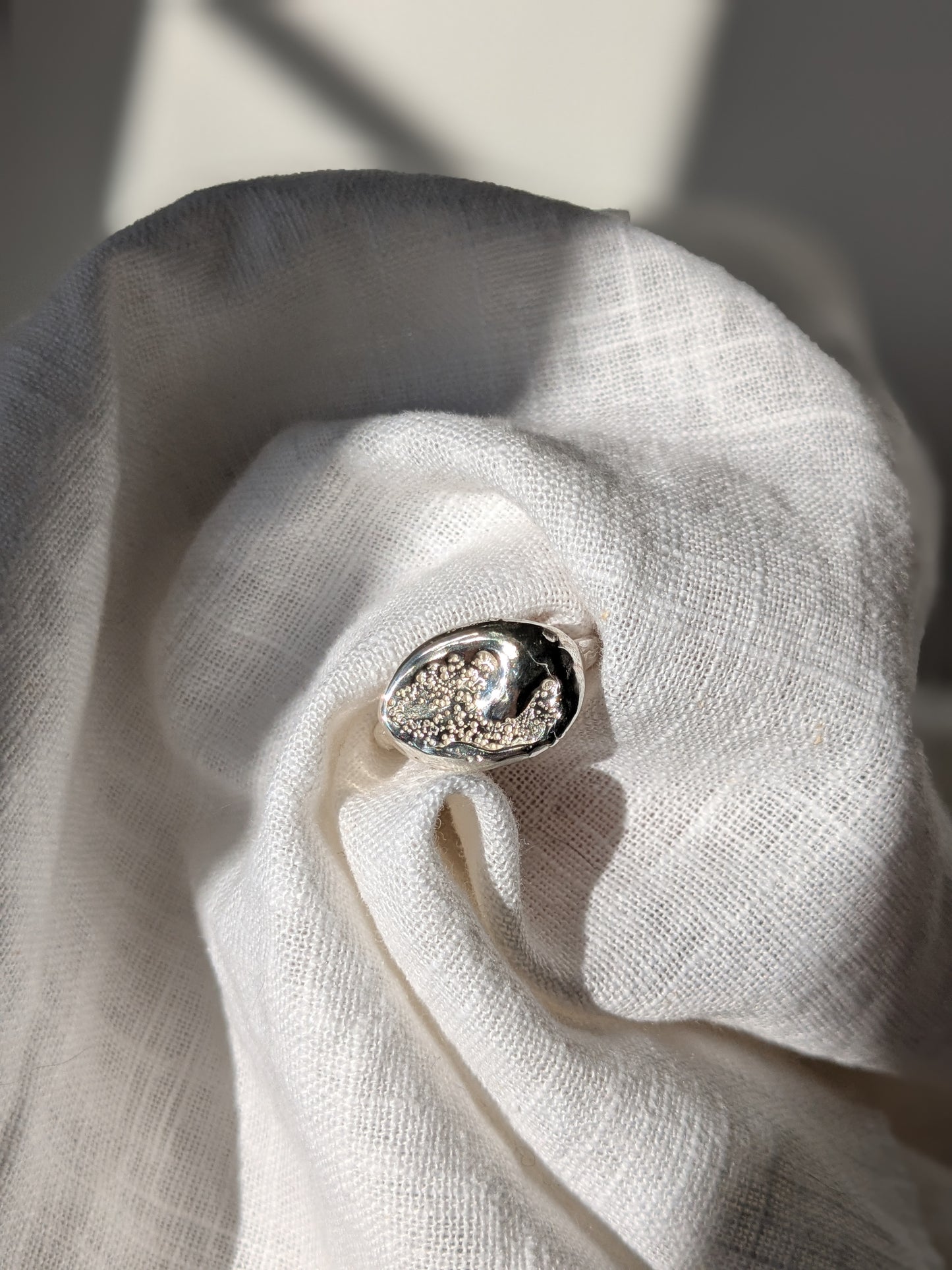 A handcrafted molten earth silver ring with an organic oval shape and textured surface, displayed on a white cloth.