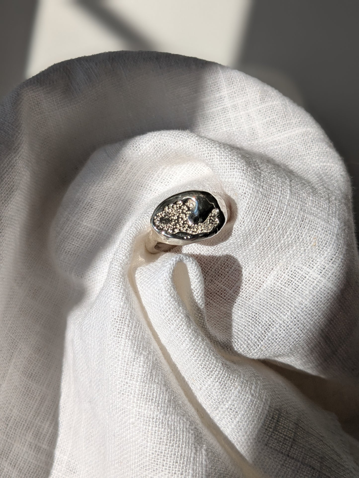 A handcrafted molten earth silver ring with an organic oval shape and textured surface, displayed on a white cloth.