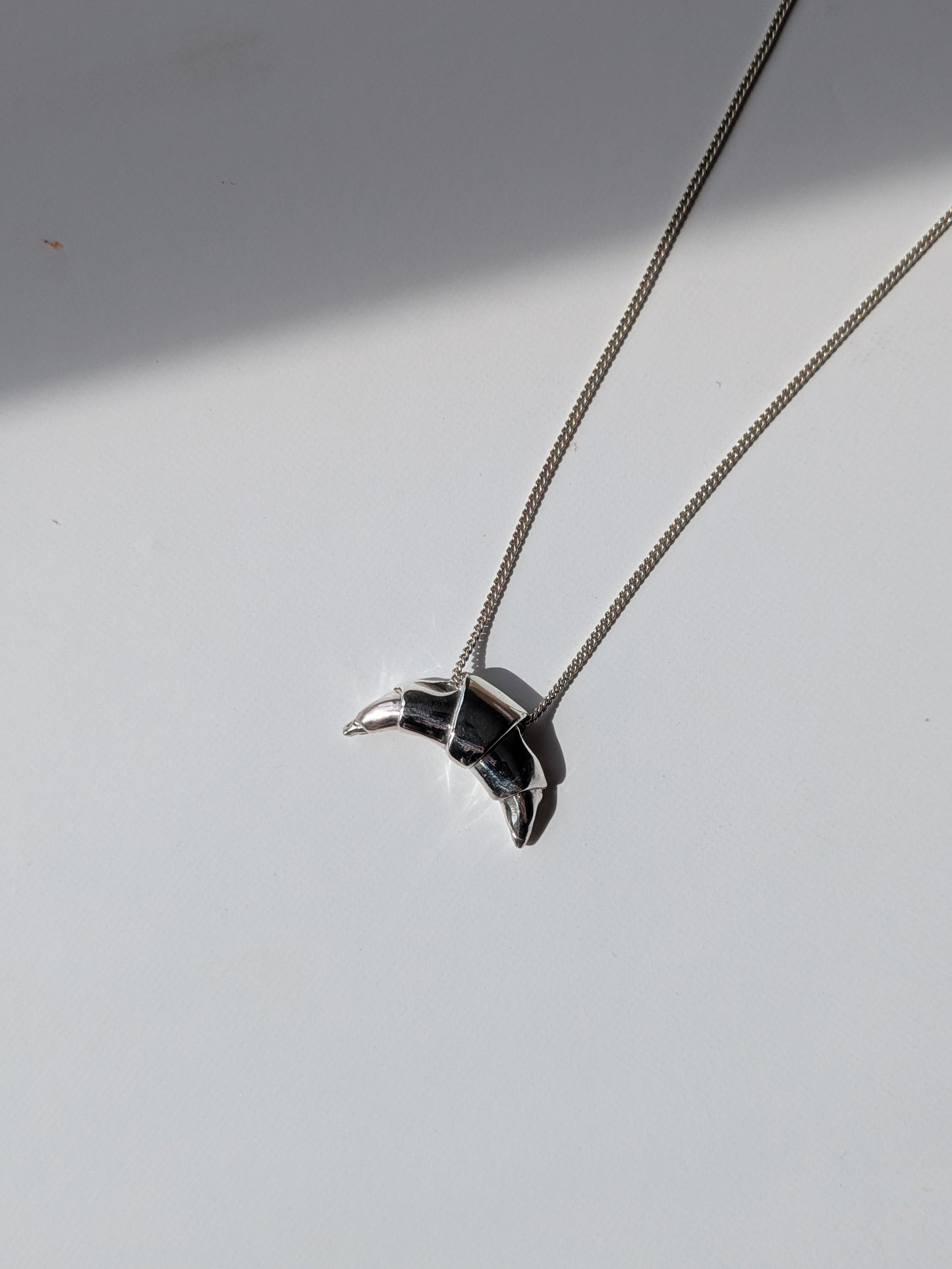 A silver croissant-shaped necklace pendant hanging on a chain, with a source of light creating a shadow and highlight on the pendant.
