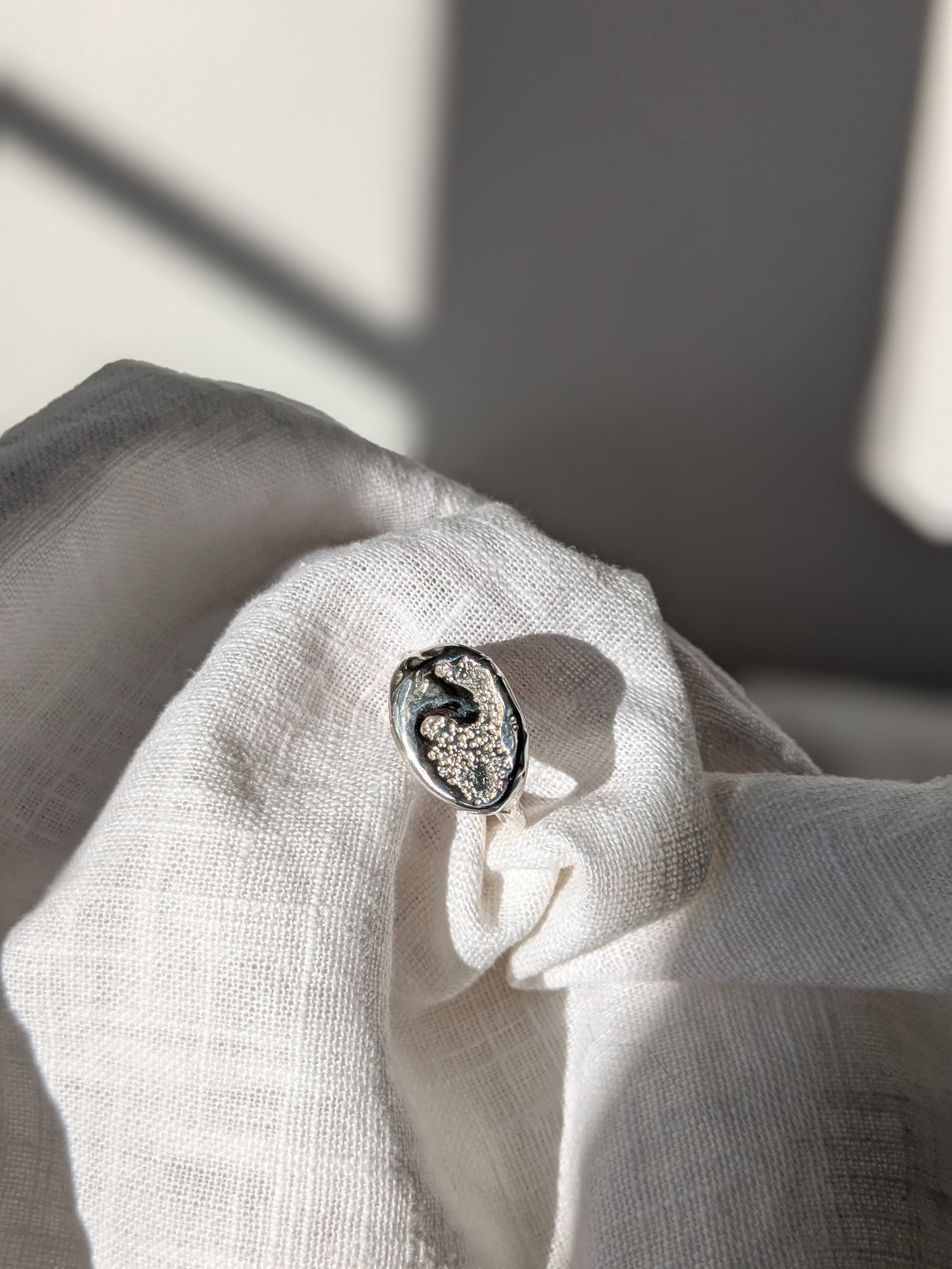 A handcrafted molten earth silver ring with an organic oval shape and textured surface, displayed on a white cloth.