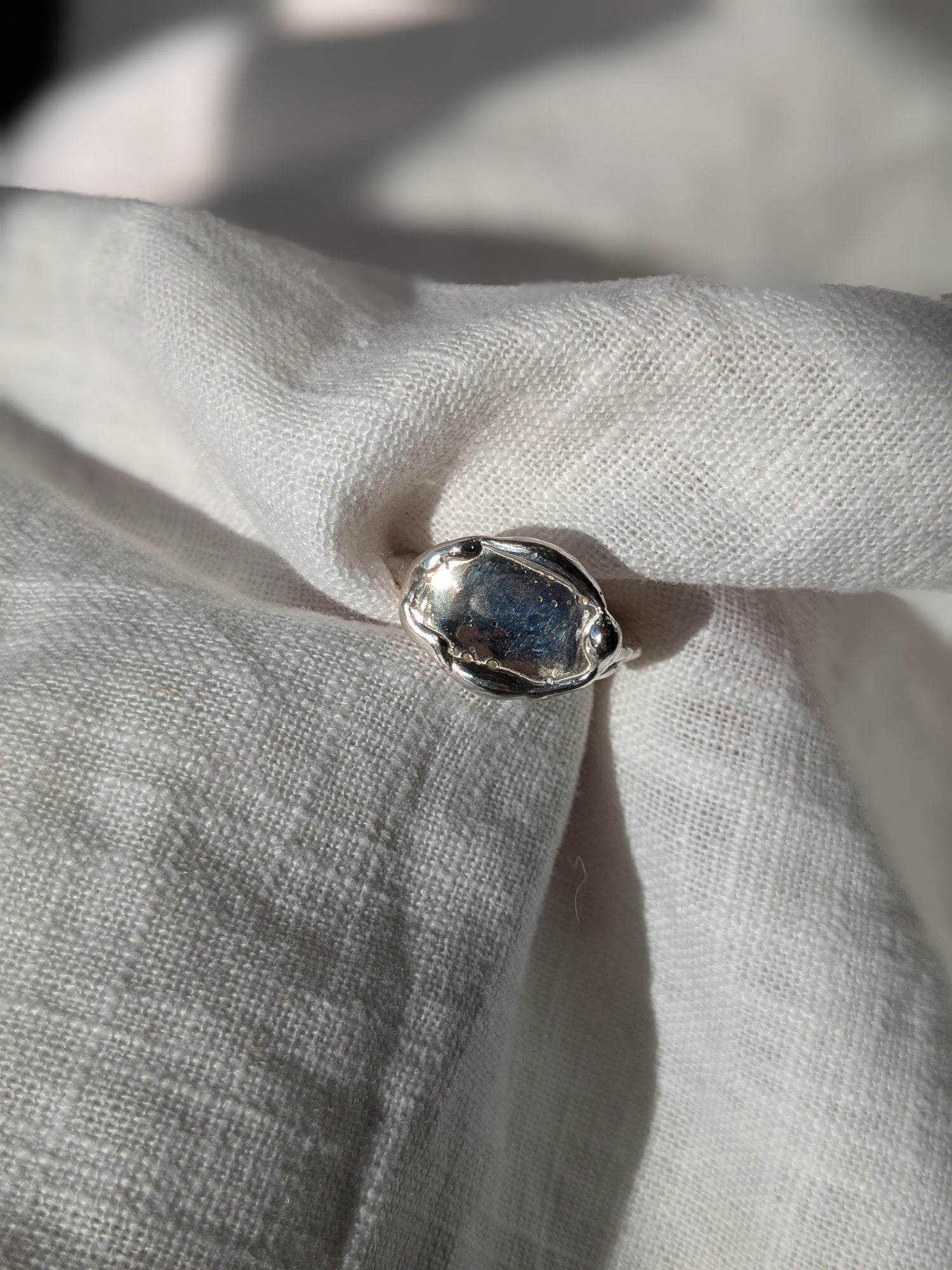 A shiny silver ring with an organic oval shape and small molten drips around the edge, placed on a textured fabric.