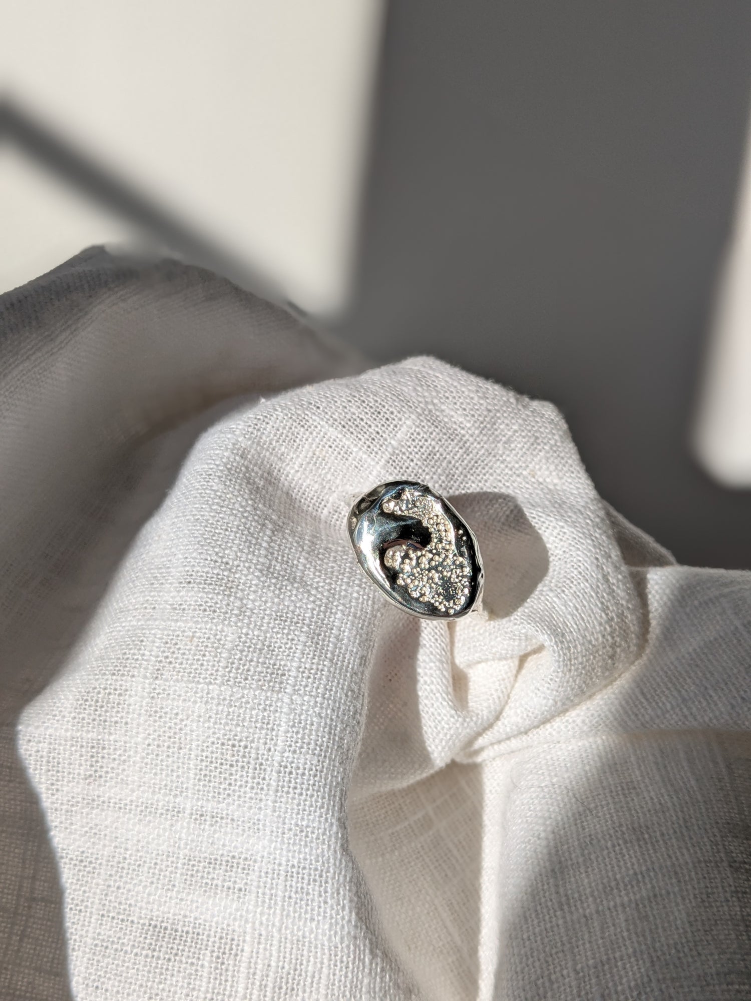 A handcrafted molten earth silver ring with an organic oval shape and textured surface, displayed on a white cloth.