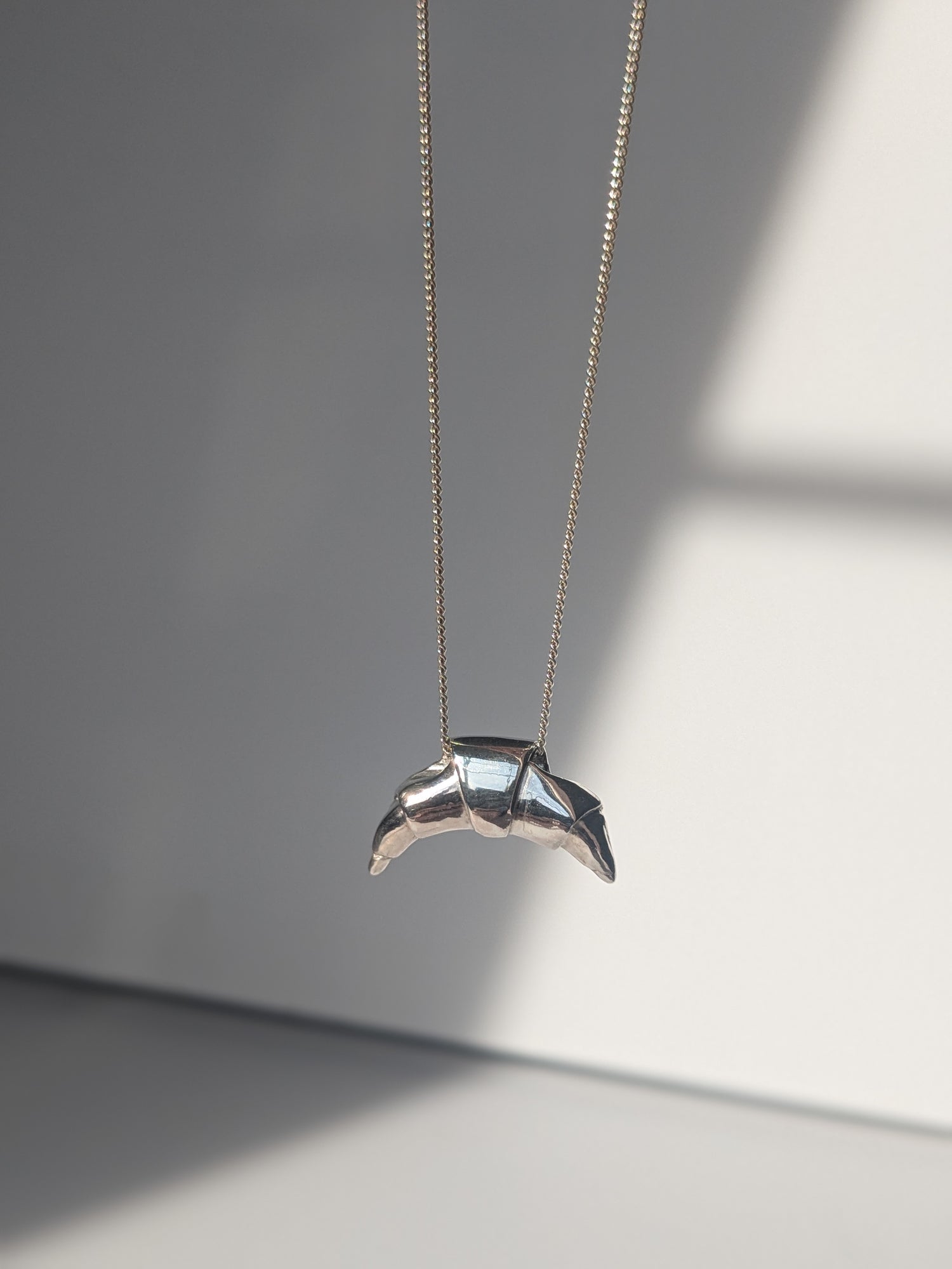 A silver croissant-shaped necklace pendant hanging on a chain, with a source of light creating a shadow and highlight on the pendant.