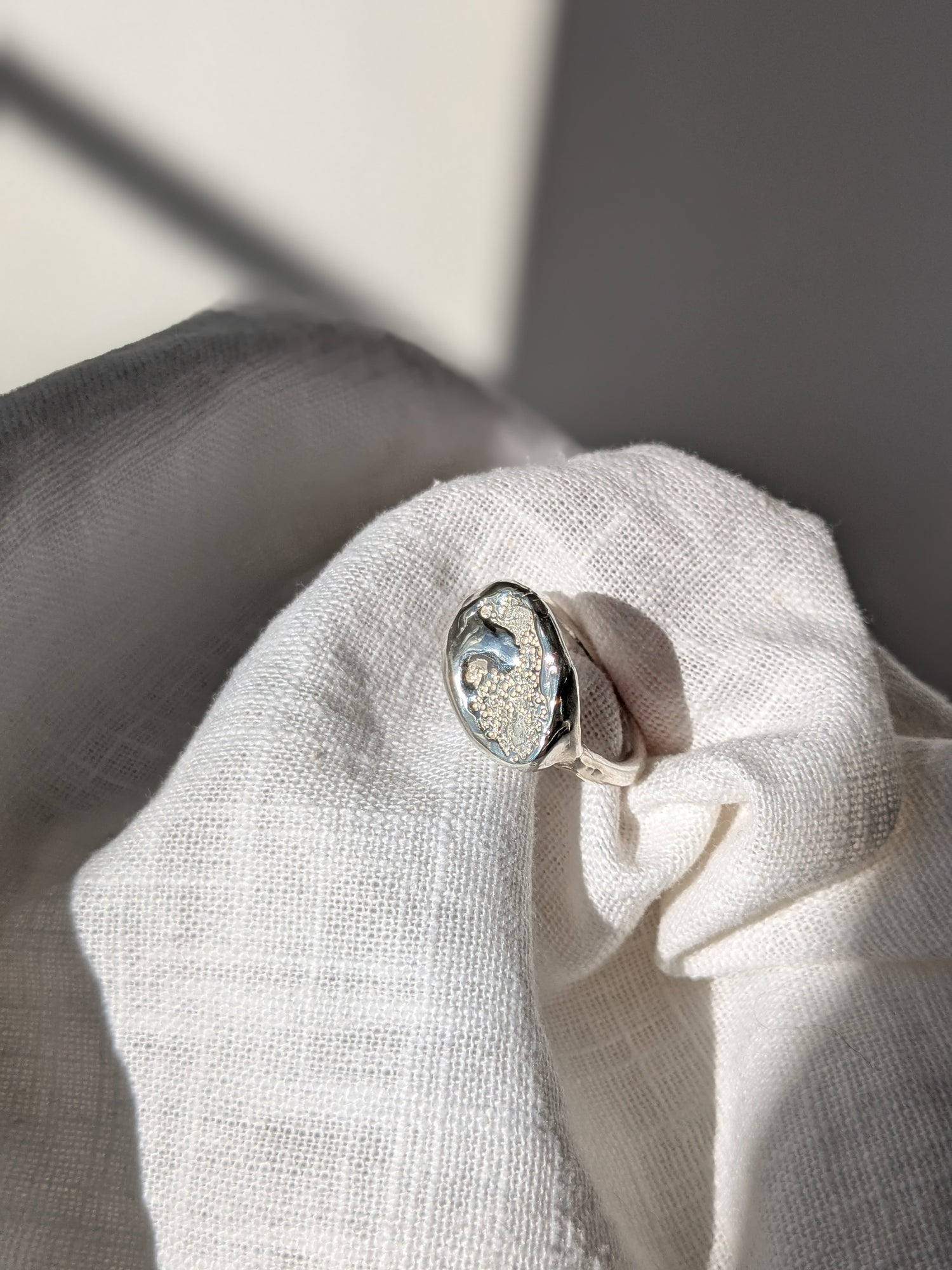 A handcrafted molten earth silver ring with an organic oval shape and textured surface, displayed on a white cloth.