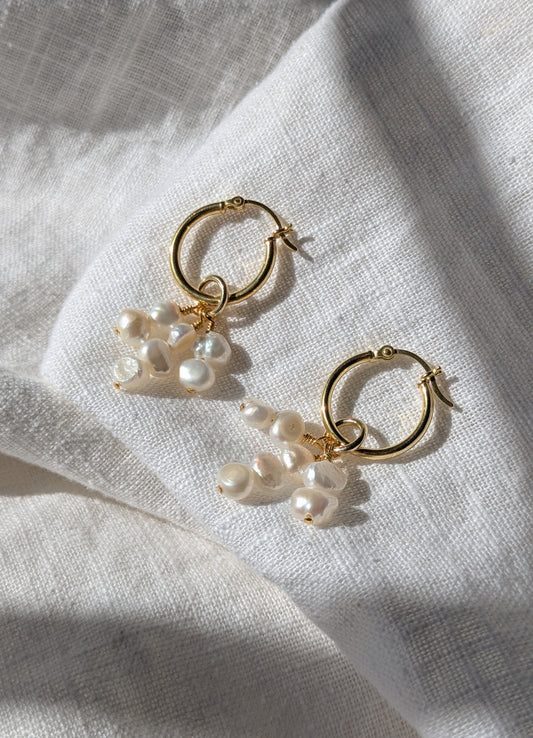 A pair of gold hoop earrings with white pearl drops, placed on a textured white fabric.