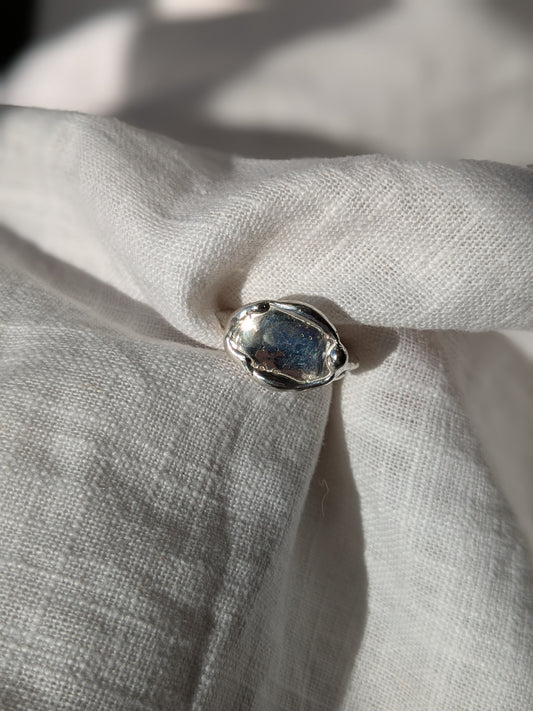 A shiny silver ring with an organic oval shape and small molten drips around the edge, placed on a textured fabric.
